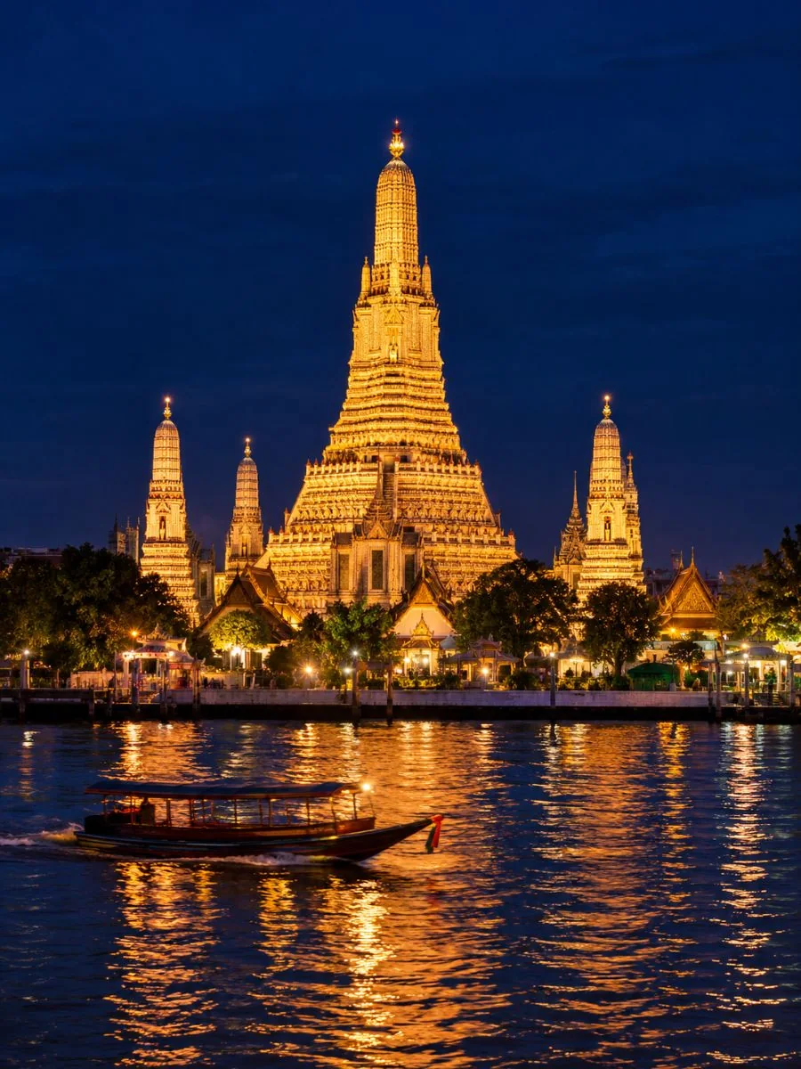 The spire of Wat Arun lit gold at night above the Chao Phraya River, seen from the opposite bank
